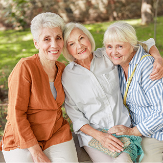 Three older women sitting outdoors on a bench, smiling and embracing each other, with greenery and trees in the background.