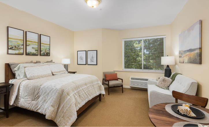 A cozy bedroom with a neatly made bed, light bedding, bedside tables with lamps, wall art, a window with blinds, an armchair, a small sofa, and a round table set with plates and a napkin.