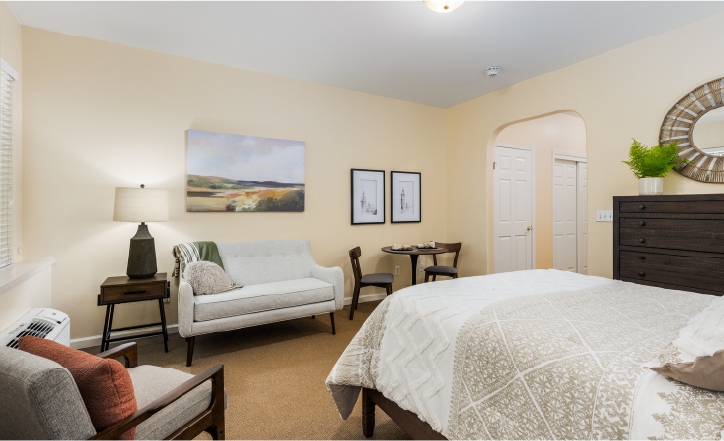 A cozy bedroom with a bed, white loveseat, small round dining table with two chairs, lamp, dresser with a mirror, and artwork on pale walls. A window lets in natural light.