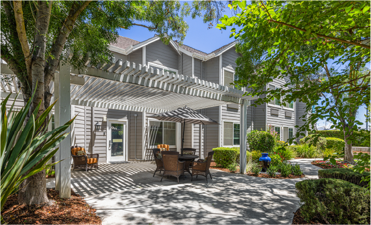 A patio with outdoor seating and a pergola is shaded by trees in front of a gray two-story building, surrounded by lush greenery and landscaped gardens on a sunny day.