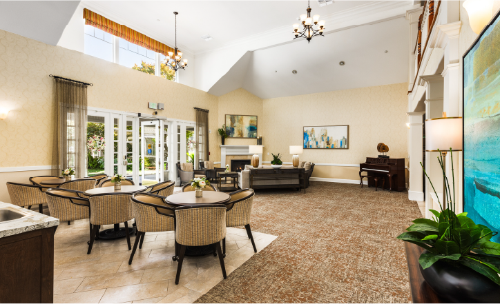 Spacious, bright lounge area with round tables and chairs, a sitting area with sofas, a piano, large windows, plants, and wall art. The room has high ceilings, chandeliers, and natural light.