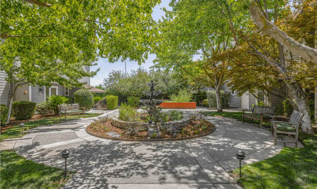 A peaceful courtyard with a circular walkway, a central black fountain surrounded by plants, benches, and trees providing shade, next to residential buildings on a sunny day.