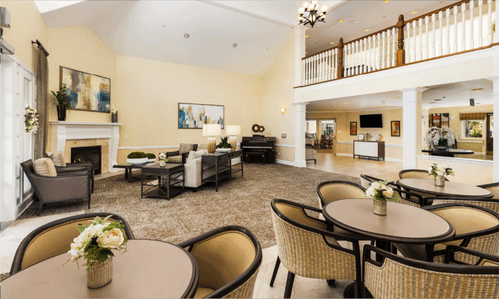 A spacious, elegant living area with round tables and cushioned chairs, a cozy seating area near a fireplace, modern artwork, and a balcony overlooking the bright, inviting room.