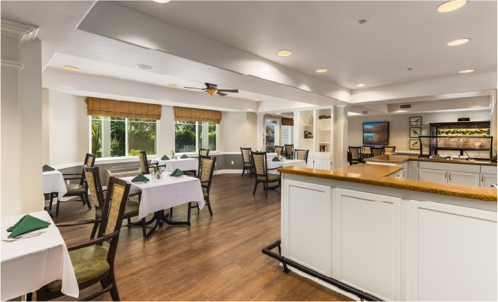 A bright, modern dining area with wooden floors, white walls, several tables set with green napkins, cushioned chairs, large windows with blind shades, and framed artwork on the walls.