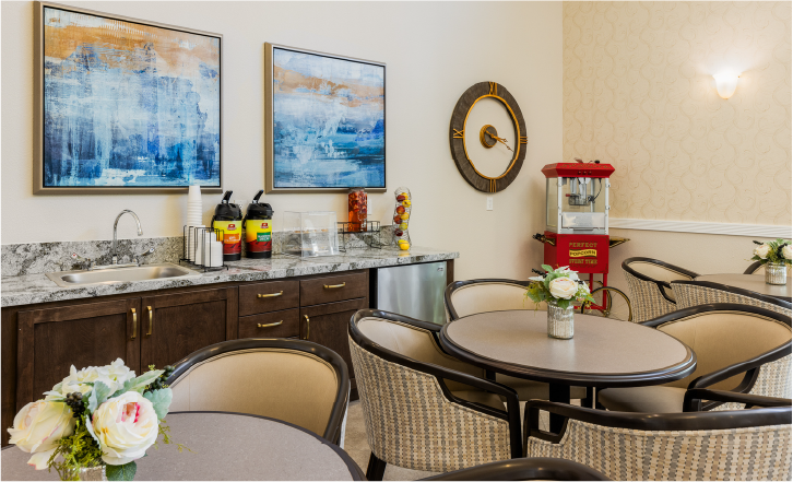 A small lounge area with round tables and chairs, floral centerpieces, a countertop with coffee dispensers, a sink, a popcorn machine, wall clock, and two abstract blue paintings on the wall.