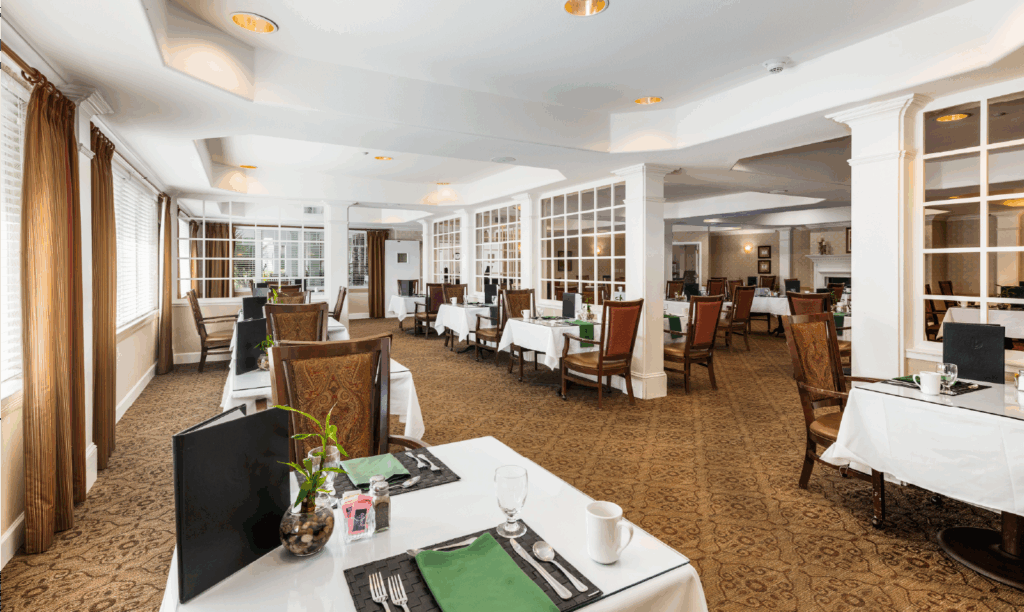 A spacious, elegant restaurant dining room with neatly set tables, brown chairs, white tablecloths, and large windows allowing natural light to fill the space. The atmosphere is clean, bright, and inviting.