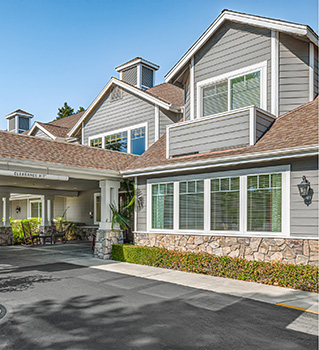 A two-story gray house with white trim and stone accents, large windows, and a covered driveway entrance, surrounded by greenery under a clear blue sky.