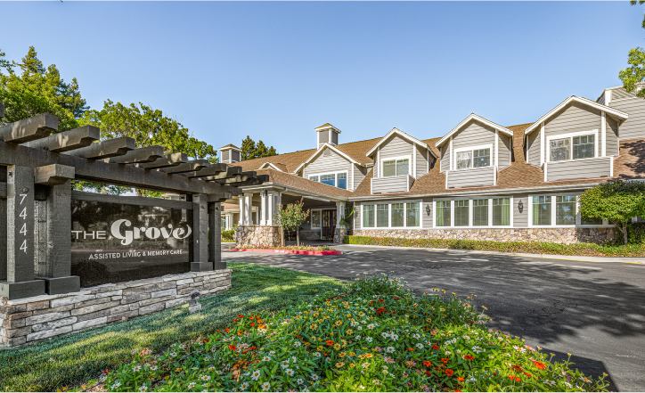 Exterior view of The Grove assisted living and memory care facility, featuring a large two-story building with brown siding, white trim, and a landscaped garden with flowers and a prominent sign at the entrance.