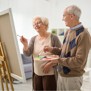 Two older adults, a woman and a man, are standing by an easel, painting together. The woman holds a brush to the canvas while the man holds a palette with colorful paints. Both appear to be enjoying the activity.