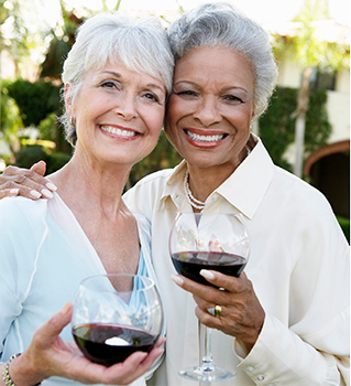 Two older women smiling and standing close together outdoors, each holding a glass of red wine. They appear happy and relaxed, enjoying each other’s company in a garden or patio setting.